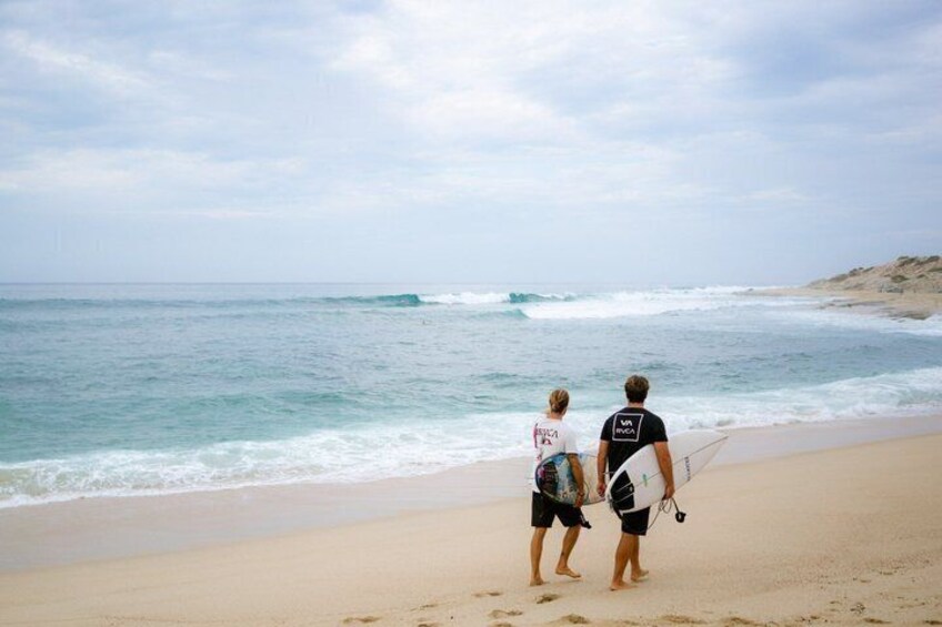 Two people walking on a beach with surfboards, ocean waves, and a clear sky. 1