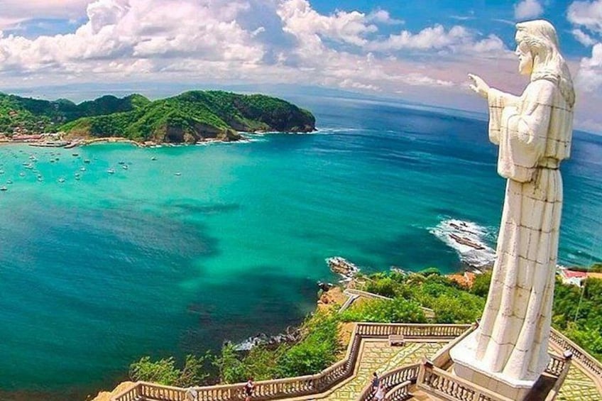 A statue overlooking turquoise waters, a coastline, and lush green hills. 4