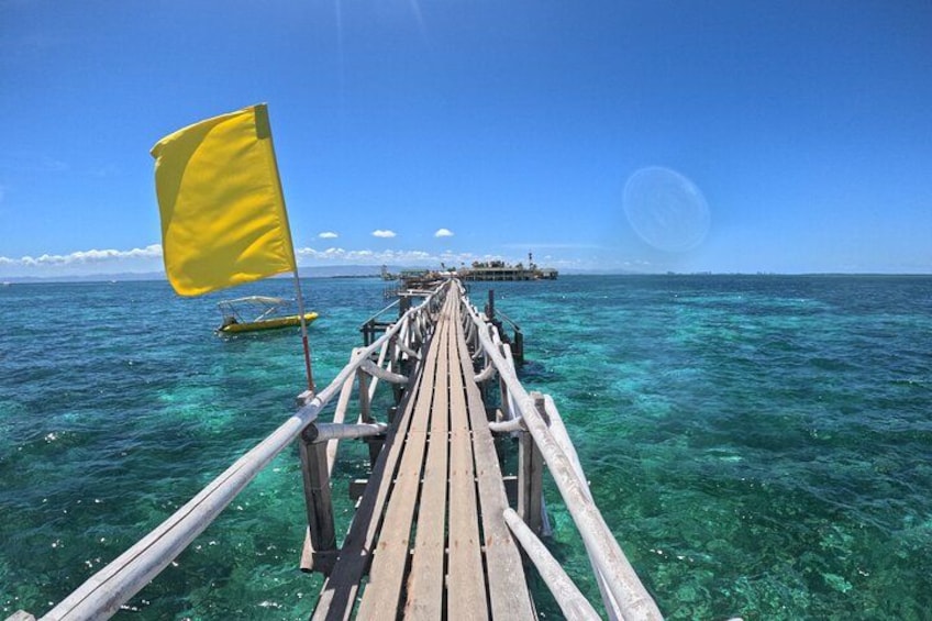 A wooden pier extending into clear turquoise water, a yellow flag, and a distant island with a structure. 5