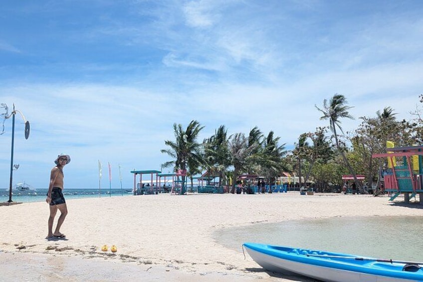 A beach with a person standing, a blue boat, palm trees, and beach huts. 4