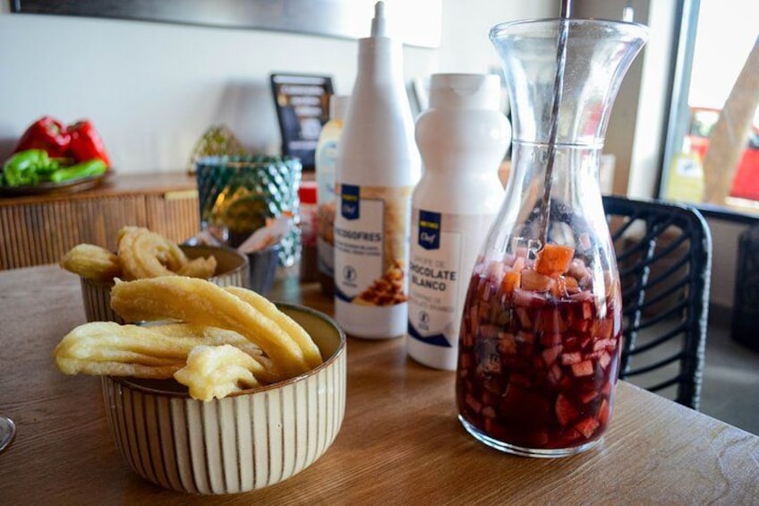 A kitchen counter with a bowl of churros, a bottle of chocolate sauce, and a glass bottle with a red liquid. 2