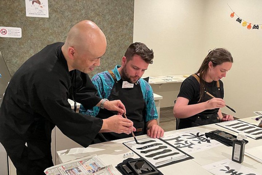 Drie personen nemen deel aan een kalligrafieworkshop, met kalligrafie-gereedschap en papier op tafel. 2