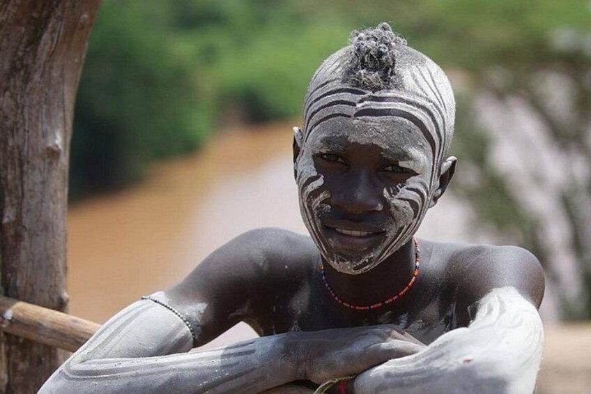 A person with traditional markings and a beaded necklace, sitting outdoors with a blurred natural background. 4