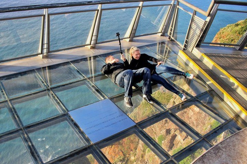 Un pont de verre avec une rambarde métallique, une personne allongée sur le pont, et une vue sur l’océan et les falaises en contrebas. 1