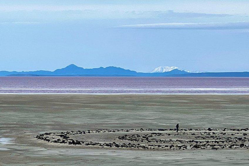A beach with a person walking, a circular stone formation, and mountains in the background. 4