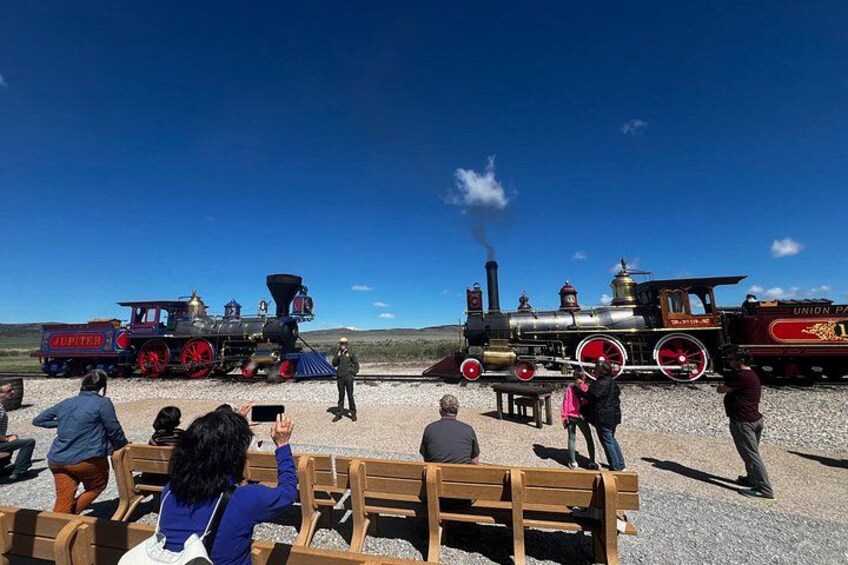 A steam train with 'JUPITER' and 'UNION PA' on the side, people sitting on benches watching, and a clear blue sky. 2
