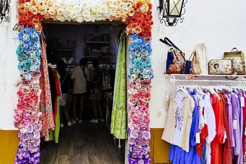 A shop entrance decorated with colorful fabric flowers, a variety of clothing items on display, and a shelf with handbags and accessories. 5