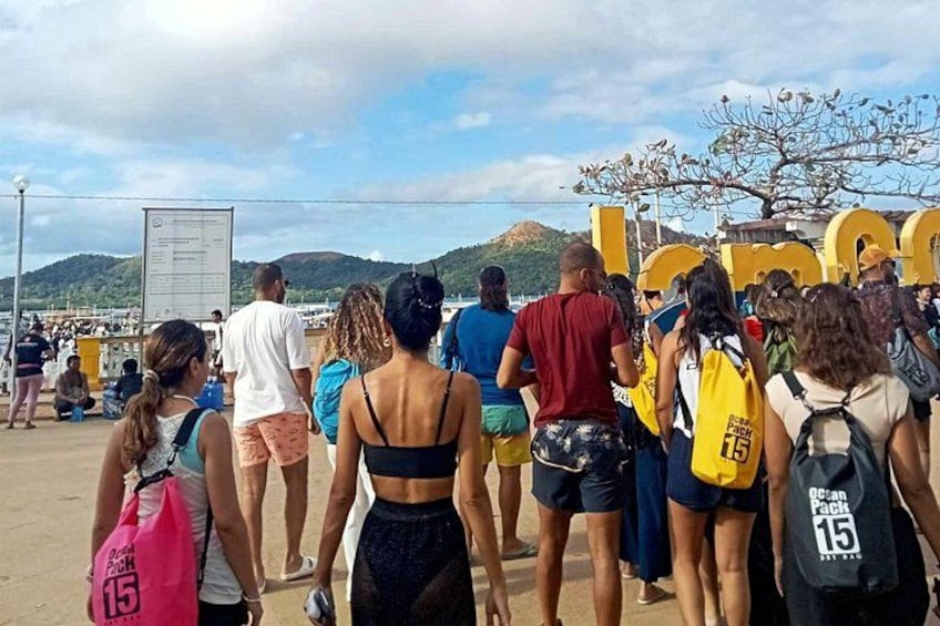 Un grupo de personas caminando por una playa, con un cartel y montañas al fondo. 2
