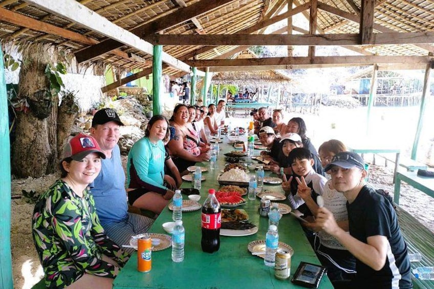 Un grupo de personas sentadas alrededor de una larga mesa al aire libre, bajo un techo de paja, con comida y bebidas sobre la mesa. 1