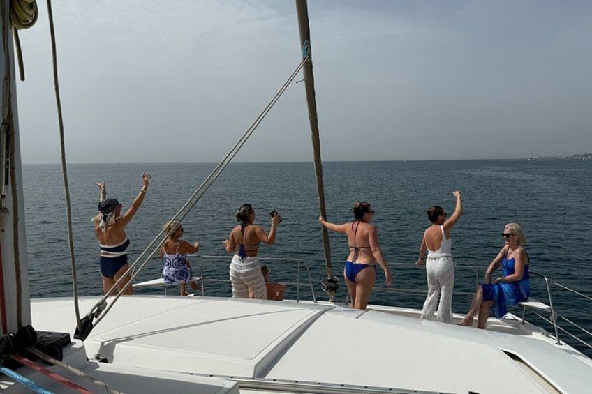 Een groep mensen op een zeilboot, sommigen staan en anderen zitten, met de zee en een heldere lucht op de achtergrond. 2