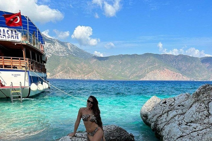A woman in a bikini sitting on rocks by a boat named 'RASAN' with a mountainous backdrop. 4