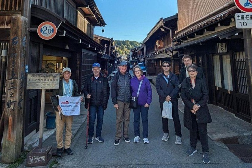 Un groupe de personnes se tient dans une rue bordée de bâtiments traditionnels en bois, avec une enseigne portant des caractères japonais et un panneau circulaire affichant un numéro. 4