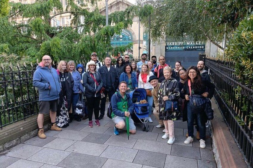 Een groep mensen poseert voor een foto in een stedelijke omgeving, met een bord waarop 'CARDIFF MALE' en 'MAREHAND CAERDYDD' staat. 1