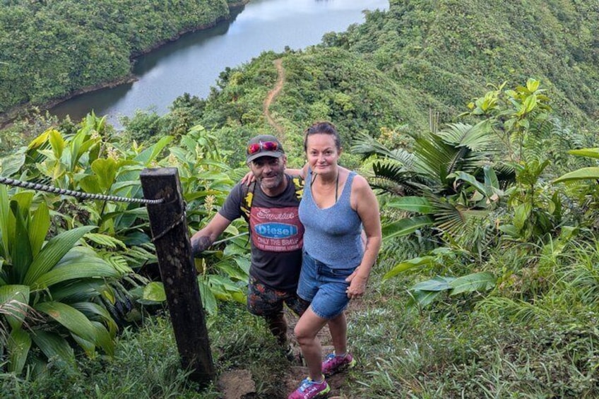 Two people standing on a hiking trail with lush greenery and a river in the background. 2
