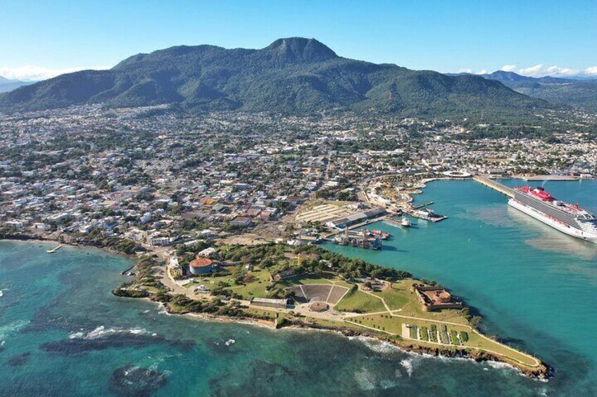 An aerial view of a coastal city with a prominent island, a large cruise ship docked at a port, and mountains in the background. 5