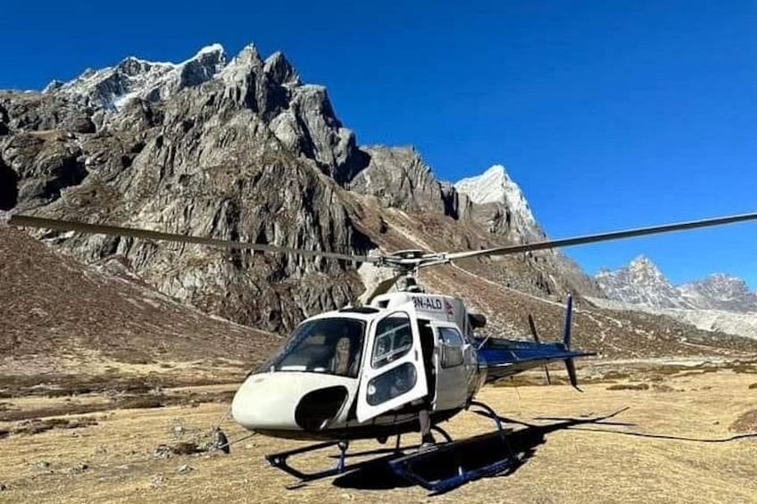 A white helicopter with 'ALTO' on the side parked on a dirt ground with a mountainous backdrop. 3