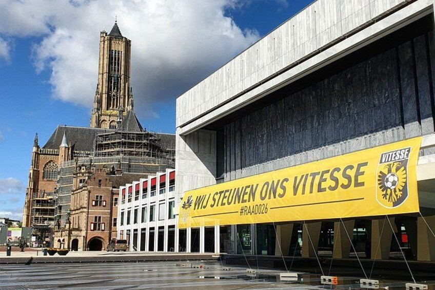 A historic church with a tall tower, a modern building with a yellow banner, and a bridge in the foreground. 1