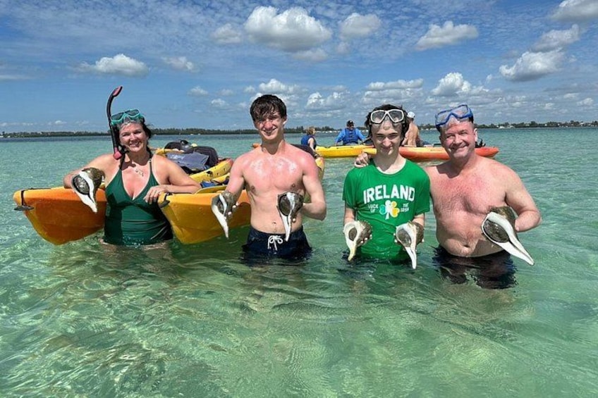 Four people in shallow water holding sea shells, with a clear sky and calm sea in the background. 1