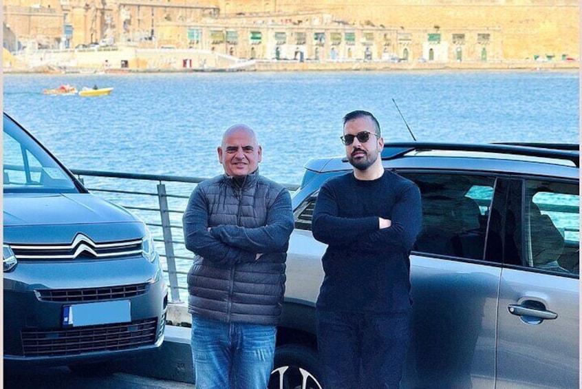 Two men standing by a car near a waterfront with buildings in the background. 5