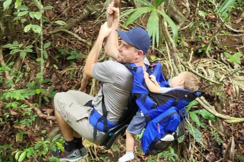 A man and a child in a forested area, both wearing blue harnesses and safety gear, with the man holding onto a rope. 2