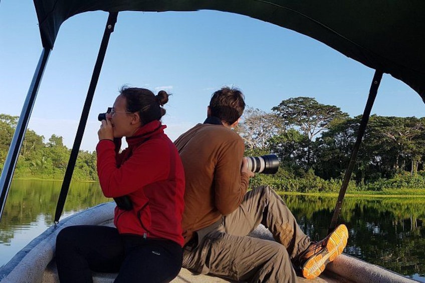 Two people on a boat, one with a camera, and a calm water body with greenery in the background. 3