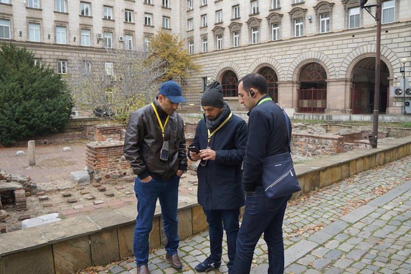 Three men standing on a cobblestone path, one holding a smartphone, with a historic building and ruins in the background. 4