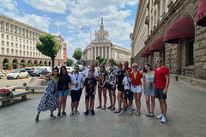 A group of people standing on a paved area with a large building in the background. 1