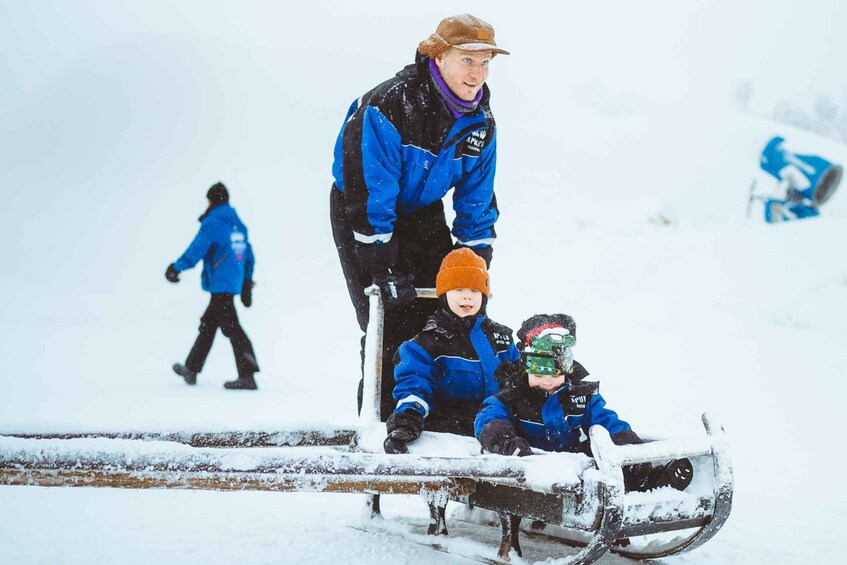 Eine Person in blauer Jacke und brauner Mütze steht auf einem Schlitten mit zwei Kindern, verschneiter Boden, und eine weitere Person geht im Hintergrund. 5