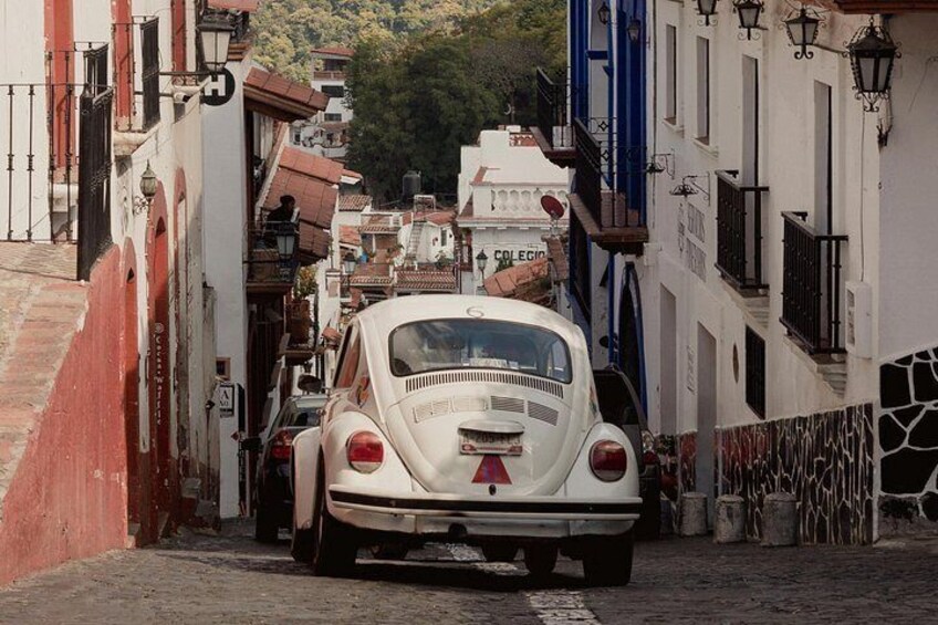 A narrow street with a vintage car parked on the side, buildings with balconies, and a sign that reads 'COLLEGIO'. 2