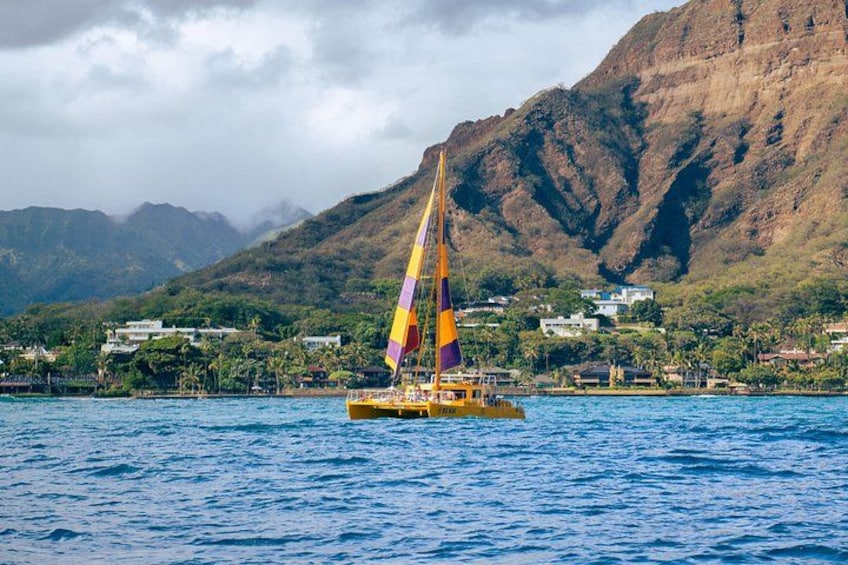 A sailboat with a colorful sail on a body of water, a mountainous coastline, and buildings in the background. 2