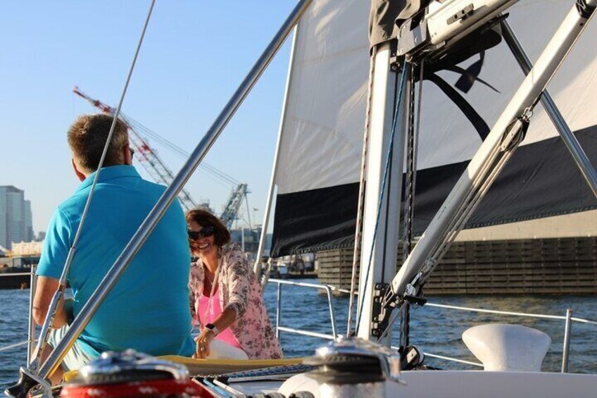 A sailboat with two people on deck, a city skyline in the background, and a crane on the shore. 5