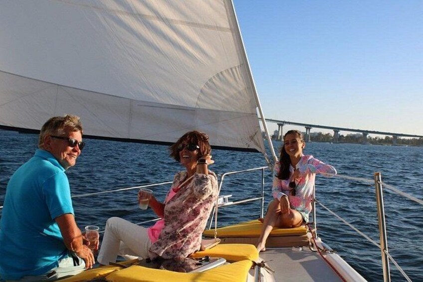 A sailboat with three people on board, a bridge in the background, and calm water. 4