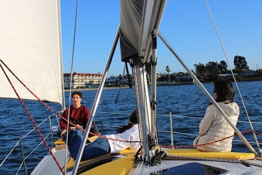 A sailboat with three people on board, one steering and two passengers, on a calm body of water with buildings in the background. 3