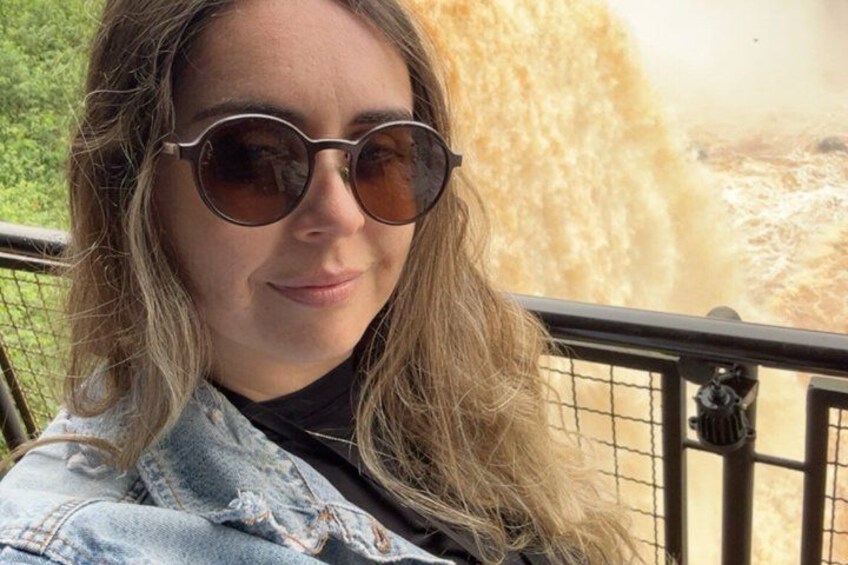 Una persona con gafas de sol y una chaqueta vaquera, de pie frente a una barandilla, con una cascada al fondo. 2