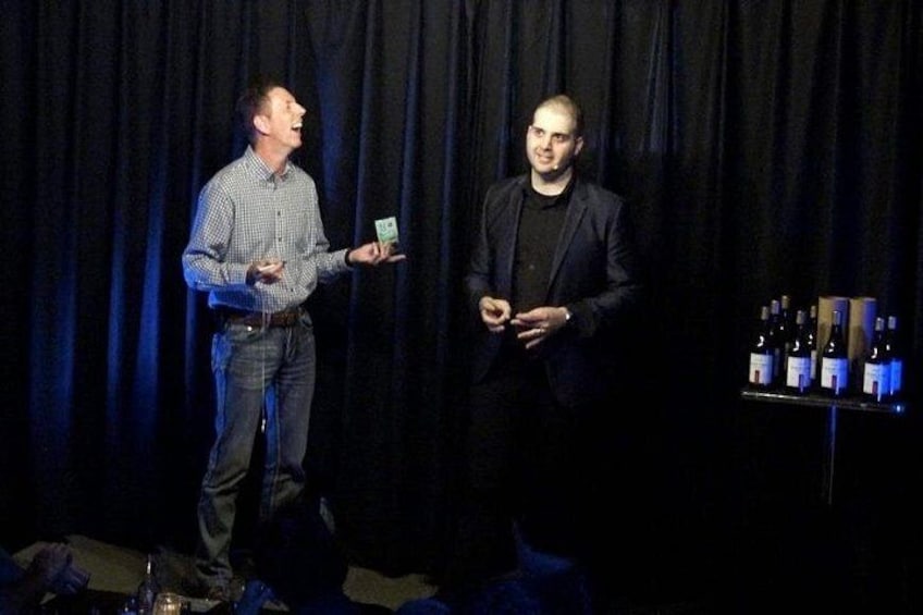 Twee mannen op een podium, met een tafel vol flessen op de achtergrond. 4