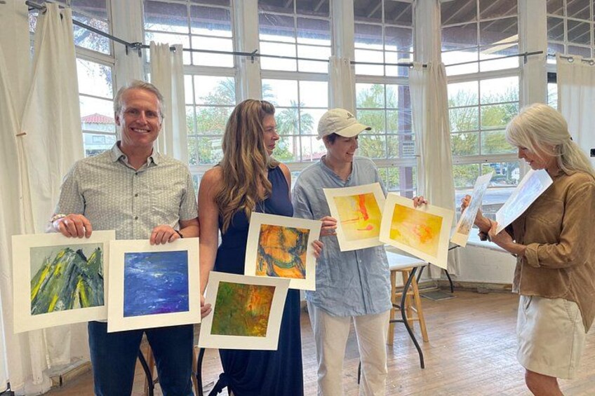 Four people holding framed artworks in a room with large windows and a wooden floor. 4