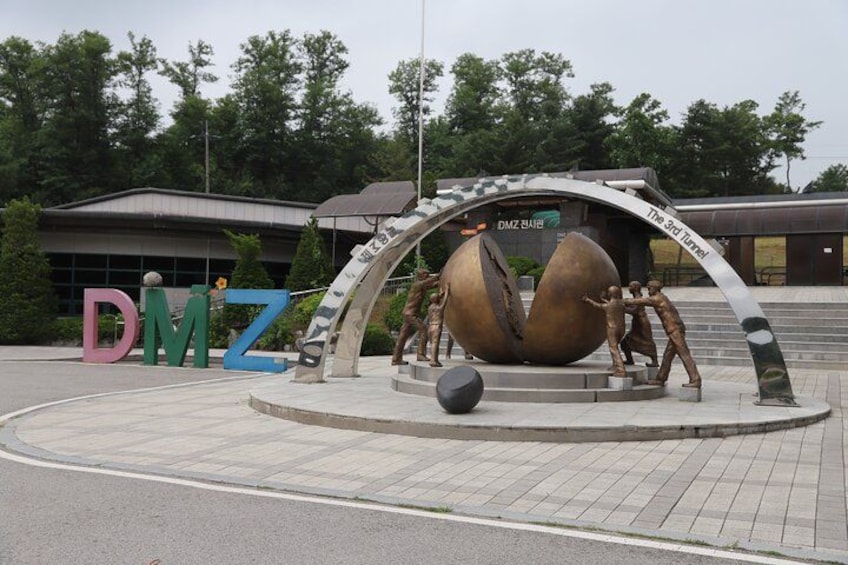 A sculpture of two large eggs with figures around them, a building with a sign reading 'DMZ,' and the letters 'DMZ' in colorful blocks. 2