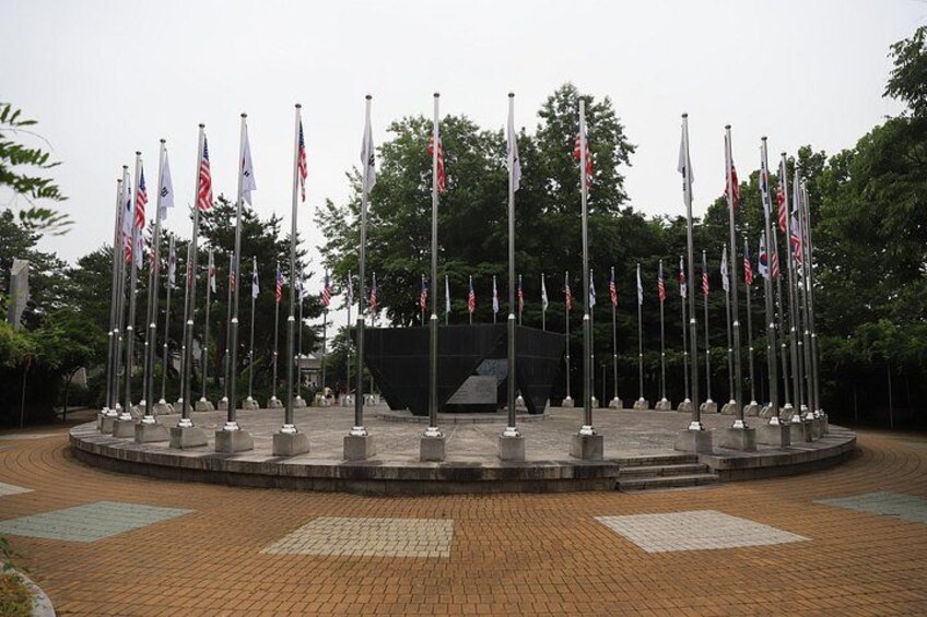 A circular plaza with multiple flagpoles, each bearing a flag, surrounded by a brick walkway and greenery. 5