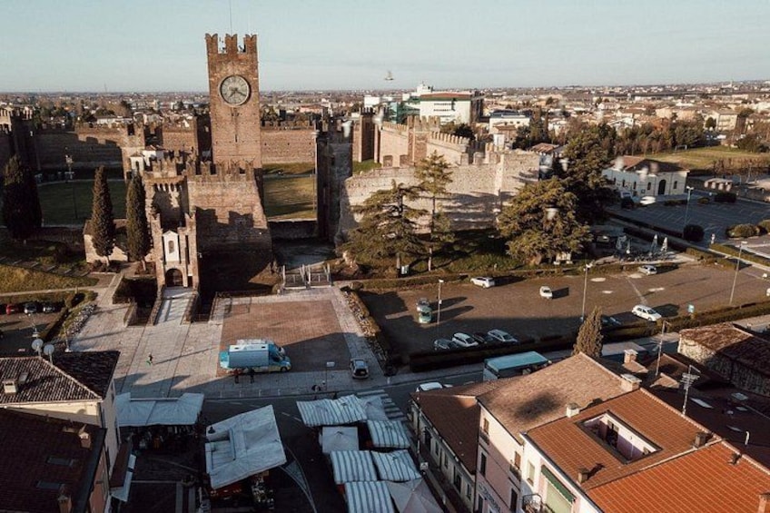 Eine historische Burg mit einem Uhrturm, umgeben von modernen Gebäuden und einem Parkplatz. 1
