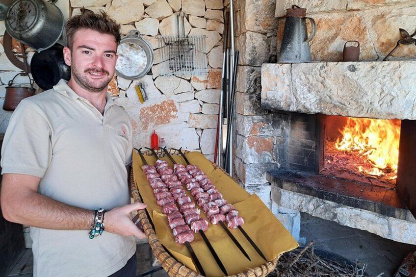 A man holding a tray of skewered meat in front of a stone fireplace with a fire burning. 5