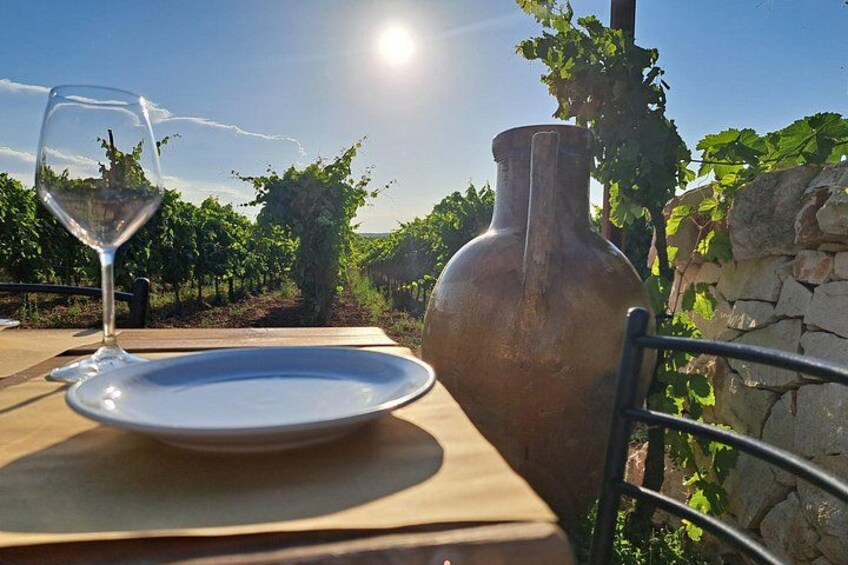 A table with a wine glass and a plate, a large ceramic vase, and a vineyard in the background. 4