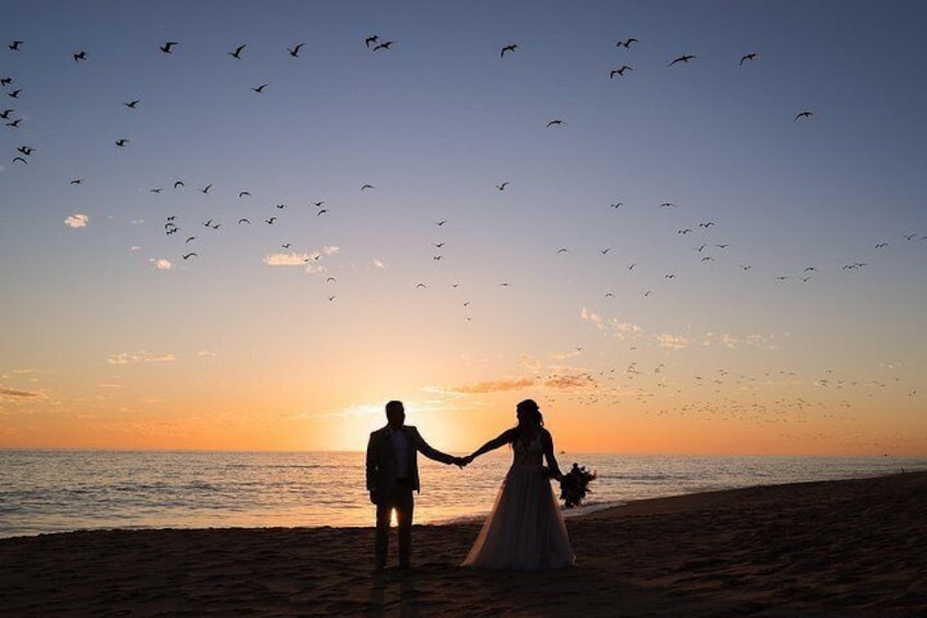 A couple holding hands on a beach at sunset with birds flying in the sky. 1