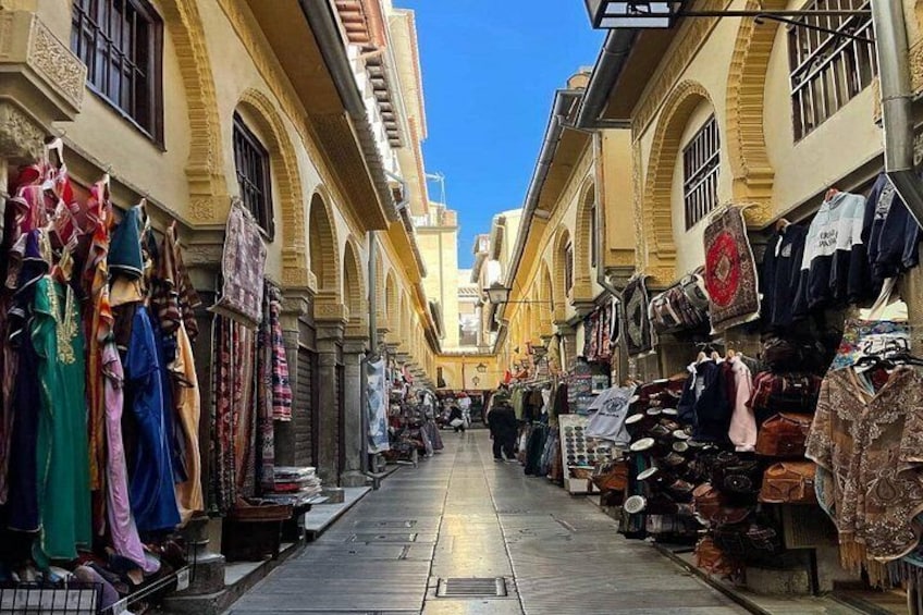 A narrow street with shops on both sides, displaying various textiles and clothing items. 1
