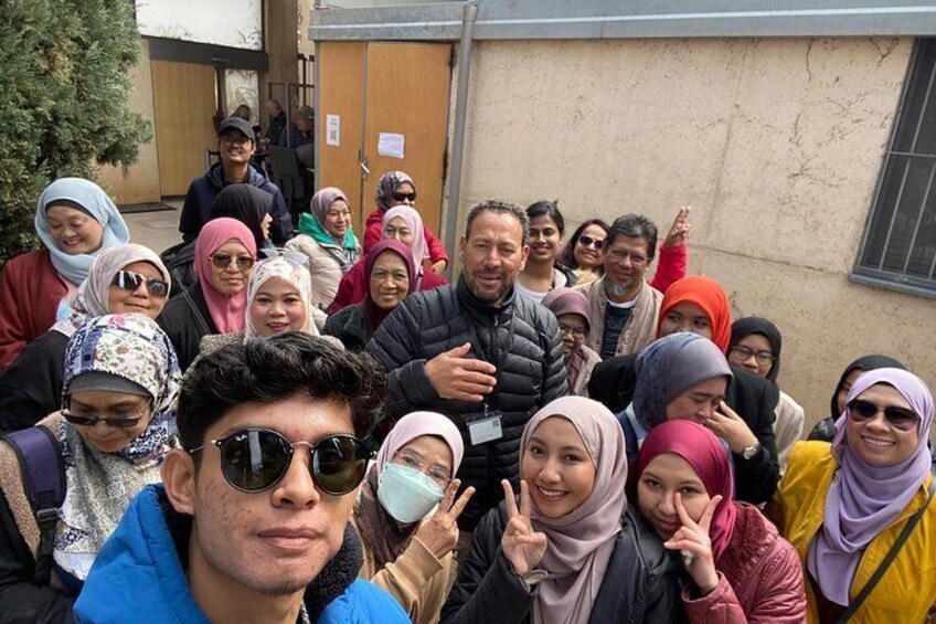 A group of people gathered outdoors, some wearing headscarves, in front of a building with a door and window. 4