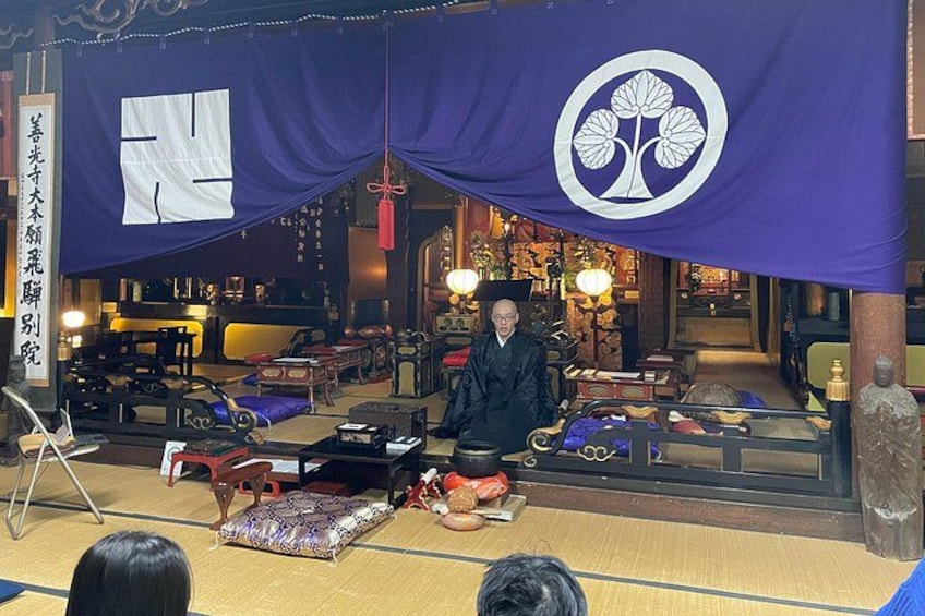 Un décor japonais traditionnel avec des tatamis, une figure assise et une grande bannière violette ornée d’un emblème blanc. 1