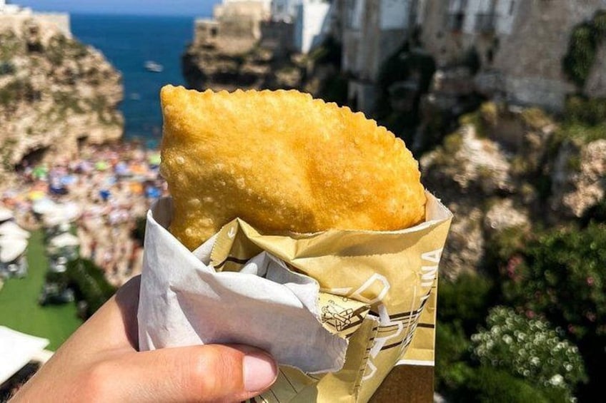 A hand holding a fried food item in a paper wrapper with a coastal landscape in the background. 3