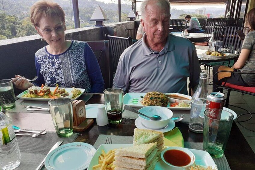 Twee personen dineren in een openluchtrestaurant met verschillende gerechten en drankjes op tafel. 1
