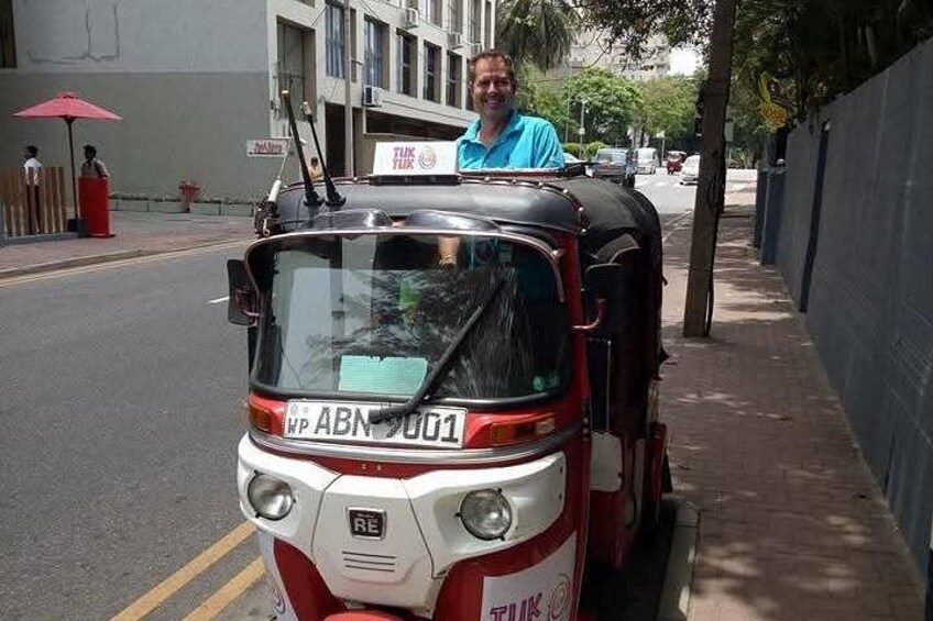 Een rode en witte tuktuk geparkeerd op een stadsstraat, met een man erachter. 3