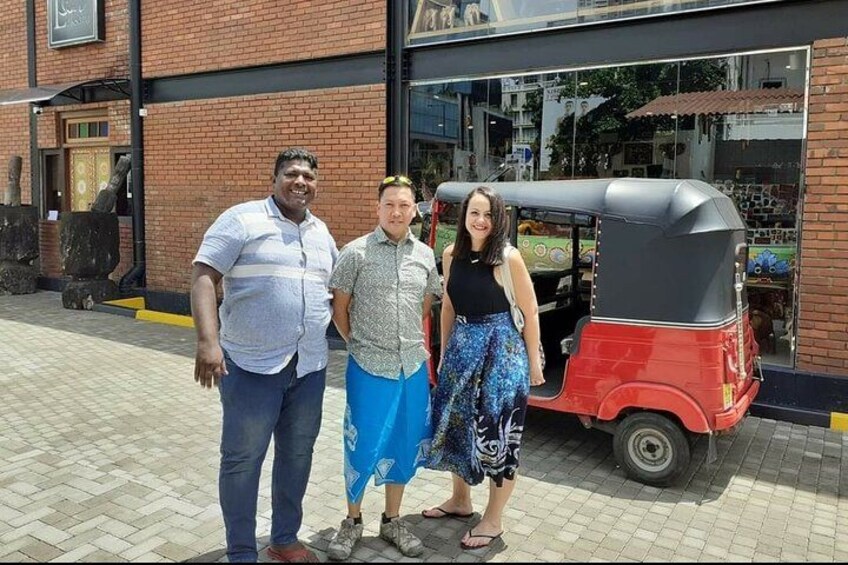 Drie personen staan voor een gebouw, met een rode tuktuk in de buurt geparkeerd. 5