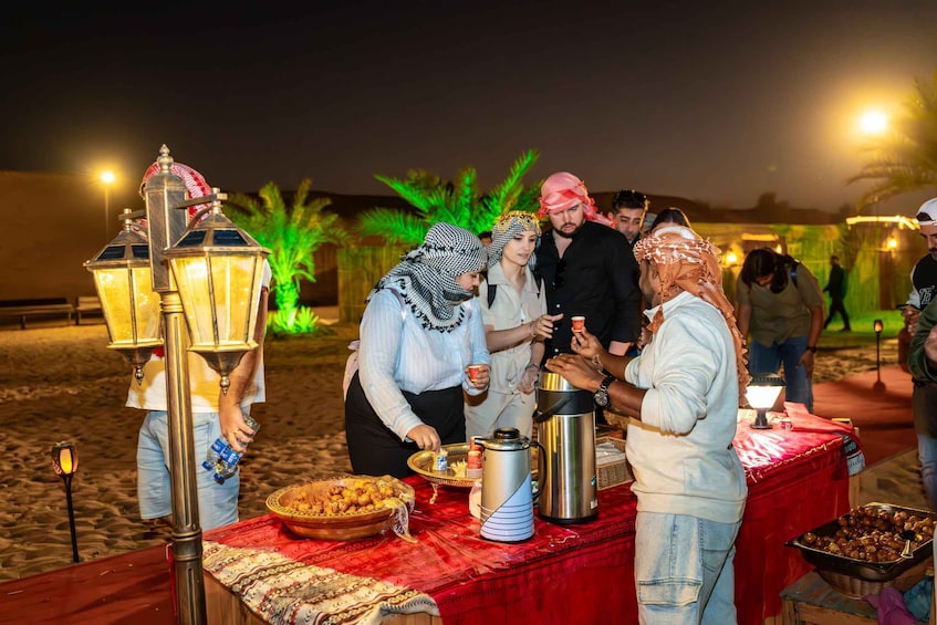 A group of people gathered around a table with food and drinks, outdoors at night with decorative lights and palm trees in the background. 3